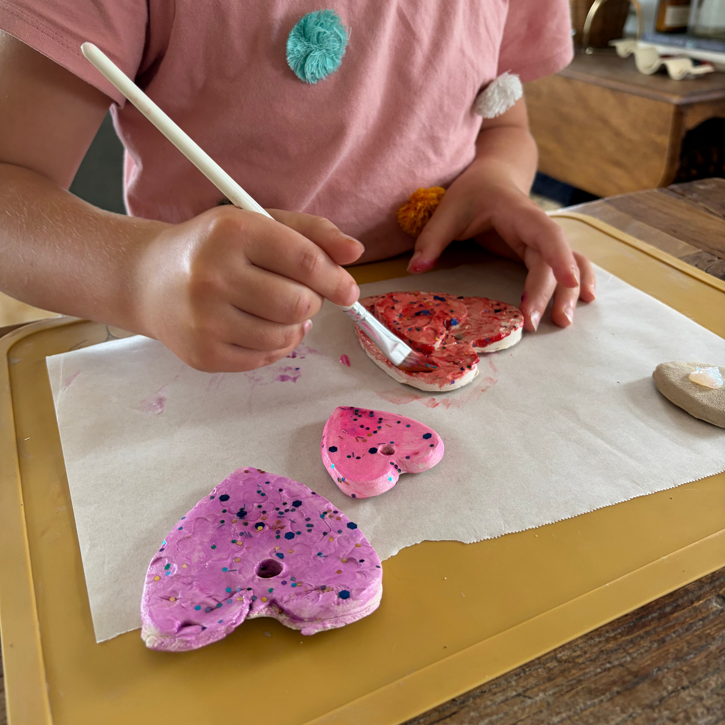 A child wearing a pink t-shirt is painting heart-shaped clay on parchment paper over a yellow tray. The clay hearts are decorated with pink, purple, and red and black sparkles.