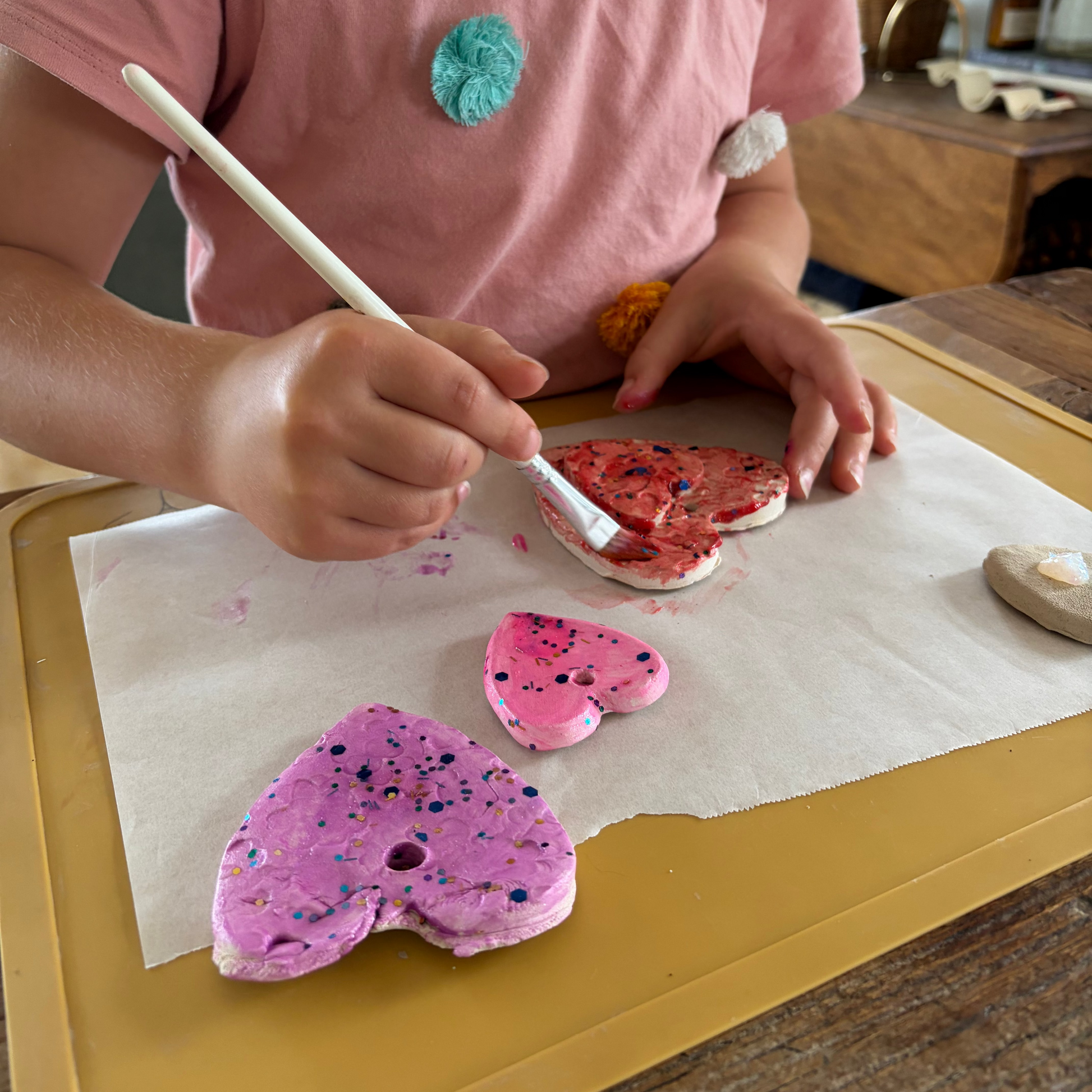A child wearing a pink t-shirt is painting heart-shaped clay on parchment paper over a yellow tray. The clay hearts are decorated with pink, purple, and red and black sparkles.
