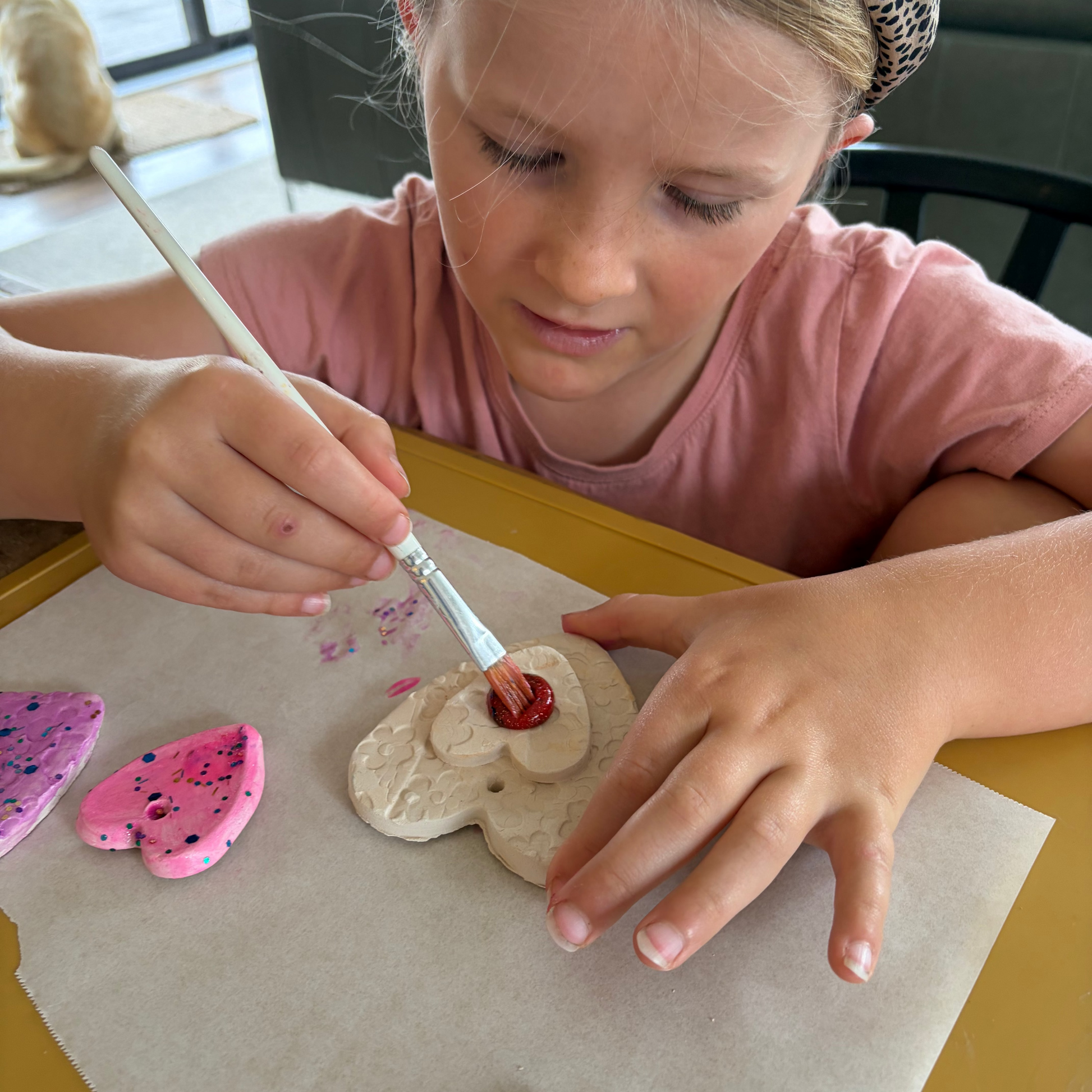 A young girl with a leopard-print headband and pink t-shirt is painting a beige clay  with a red glitter paintt on a parchment paper sheet. Pink painted clay pieces are visible on the left, 