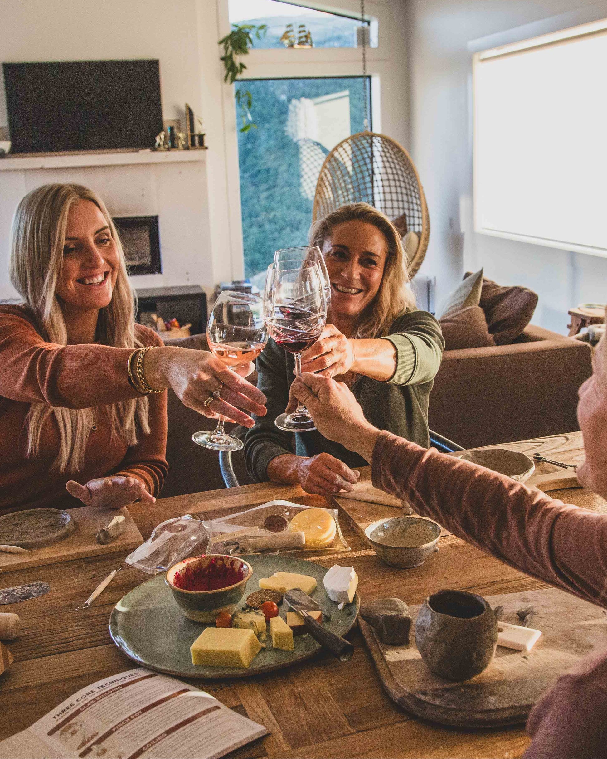 Three women toasting with wine glasses creating with clay in a cozy living room.