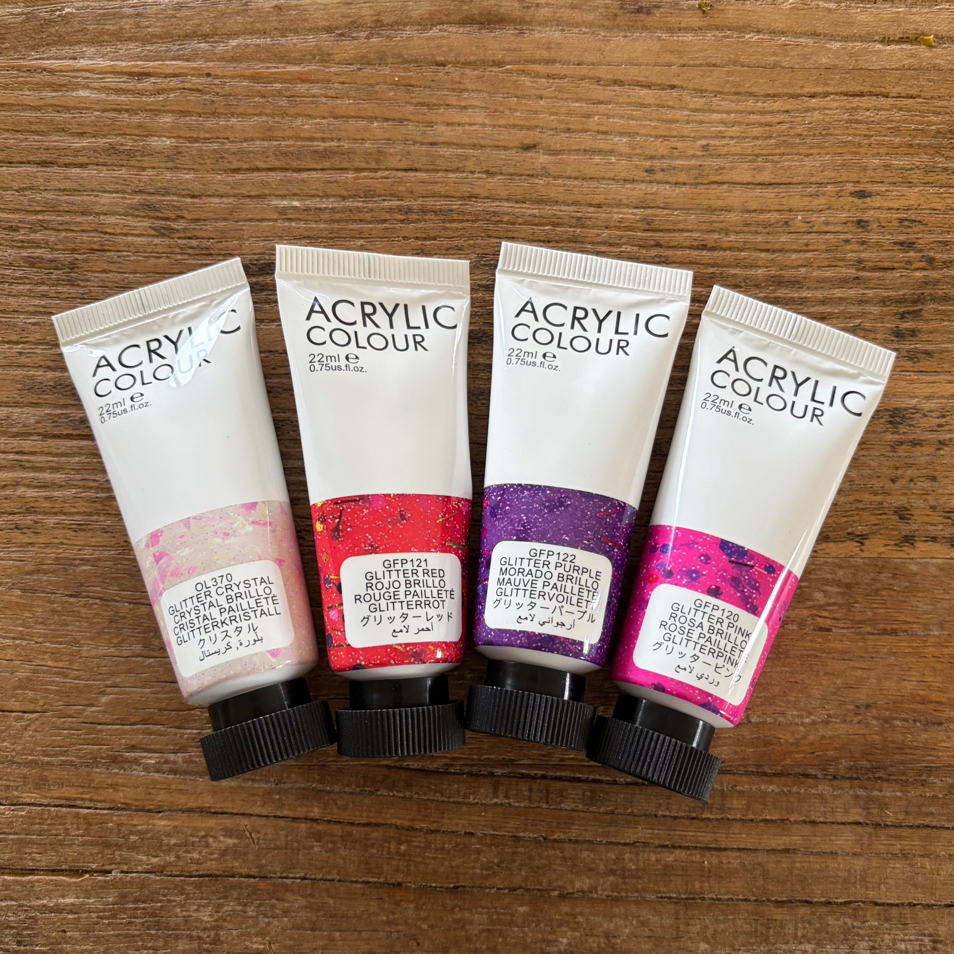 Four tubes of glitter acrylic paint in pink, red, purple, and magenta shades, arranged side-by-side on a wooden surface.