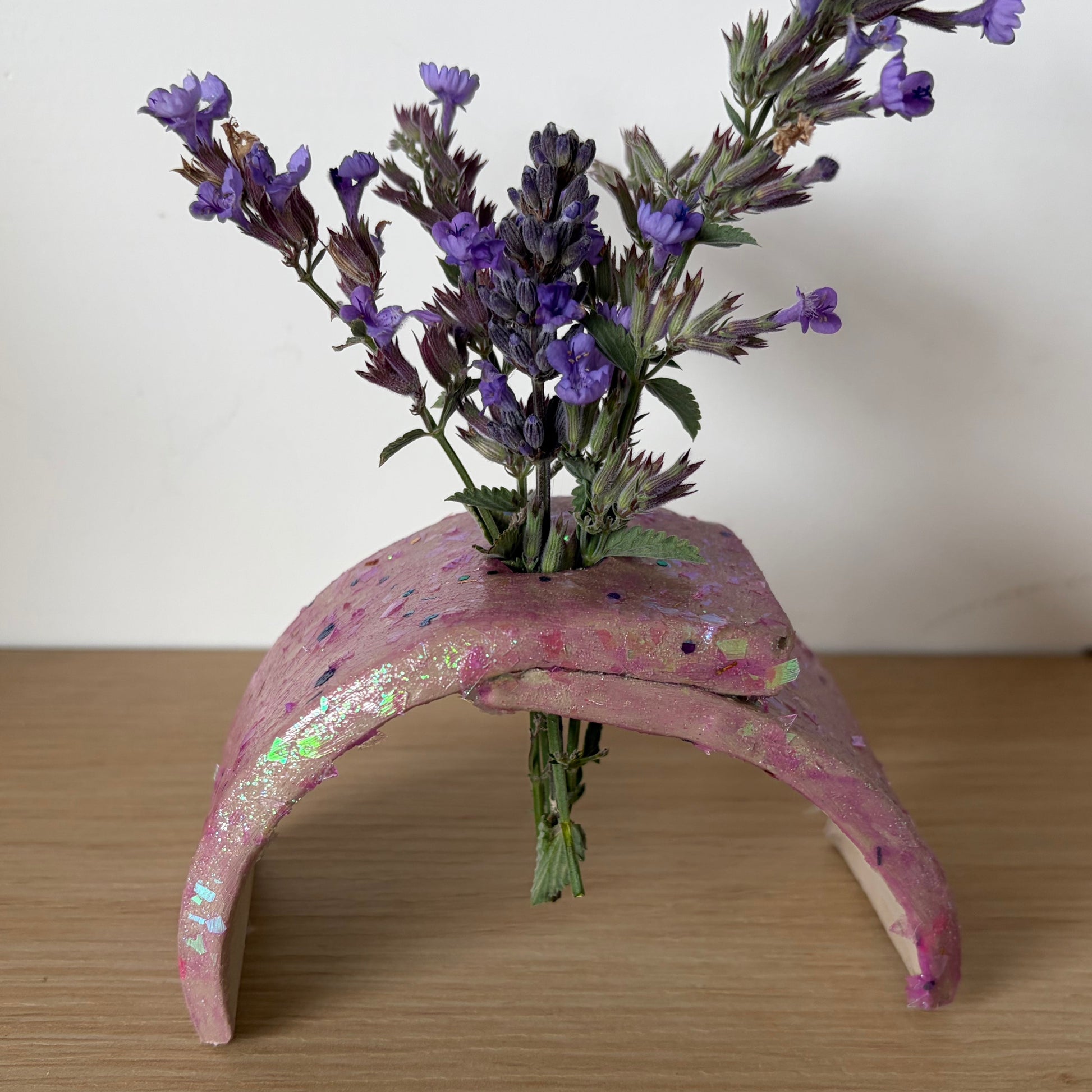 A pink ceramic vase with a glossy finish and subtle glitter , holding a small bouquet of purple lavender flowers, set on a light wood tabletop against a plain off-white wall.