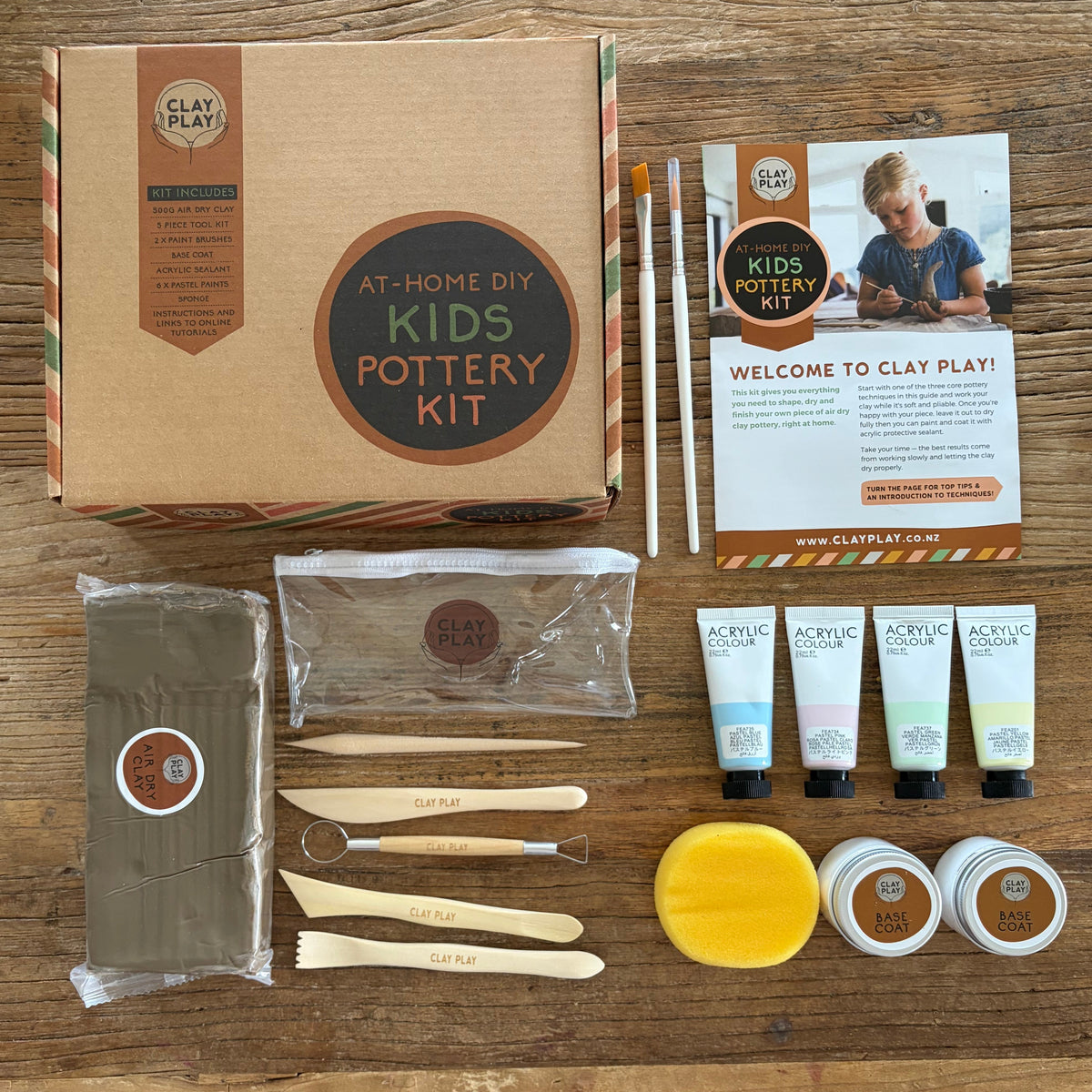 A DIY Kids Pottery Kit laid out on a wooden table, including a kraft paper box, clay, paint tubes, brushes, tools, and a welcome card. 