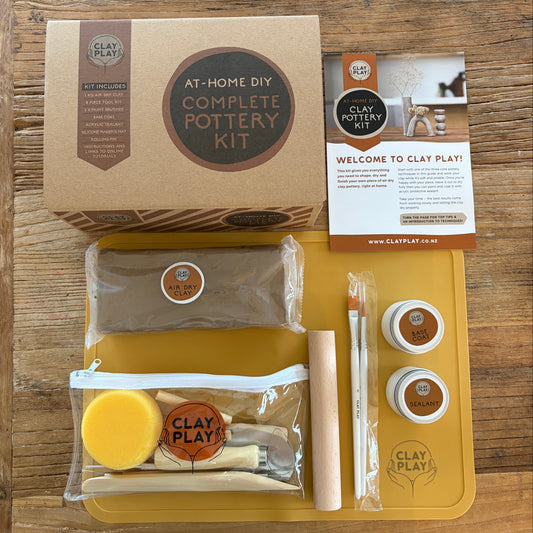 Clay Play At-Home DIY Complete Pottery Kit laid out on a wooden table, including a brown kraft box, a yellow silcone mat a sponge, tools, and a welcome card.