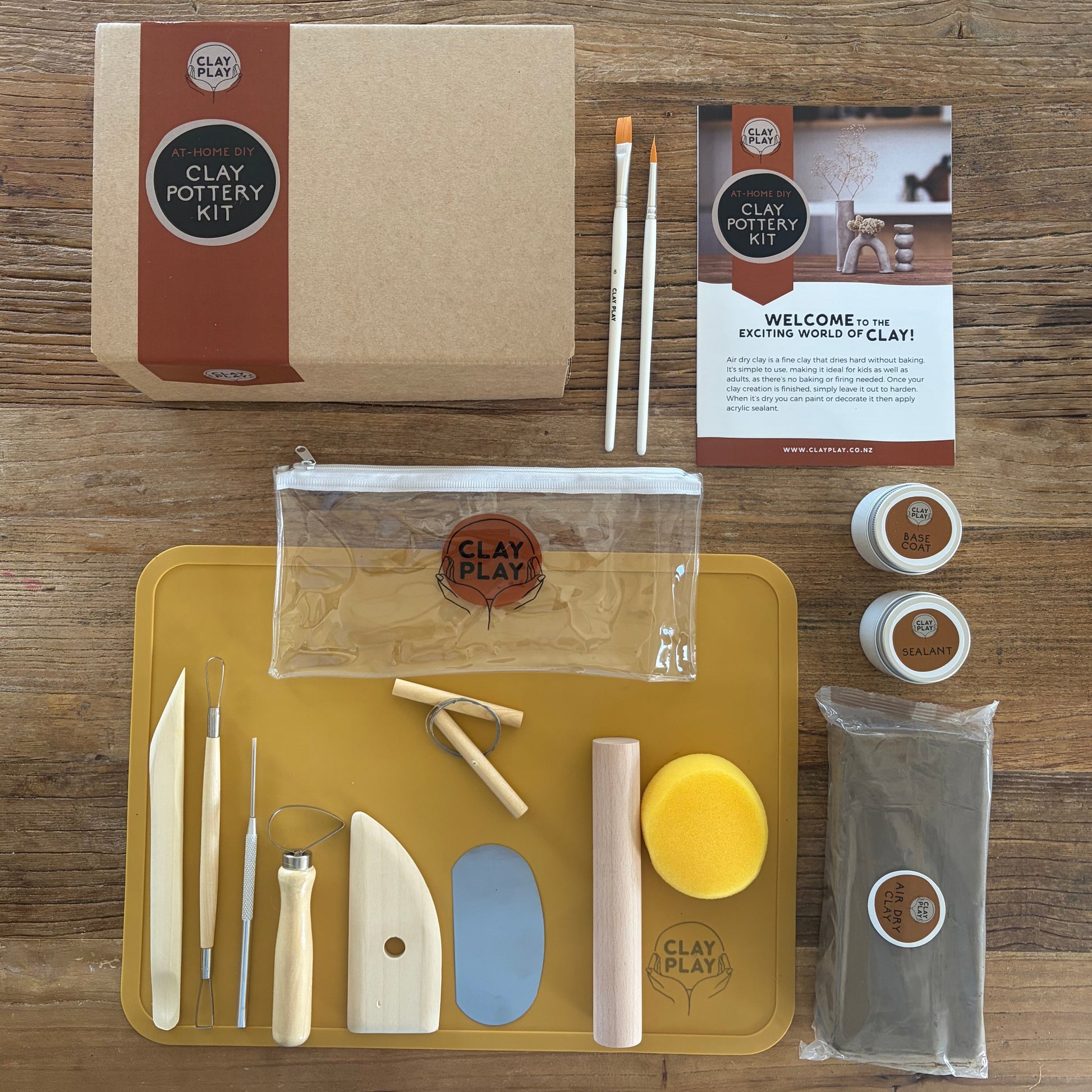 A Clay Play pottery kit laid out on a wooden table, including a kraft paper box, a yellow tool tray with sculpting tools, two small jars of clay, a bag of clay, a clear pouch, and a welcome card.