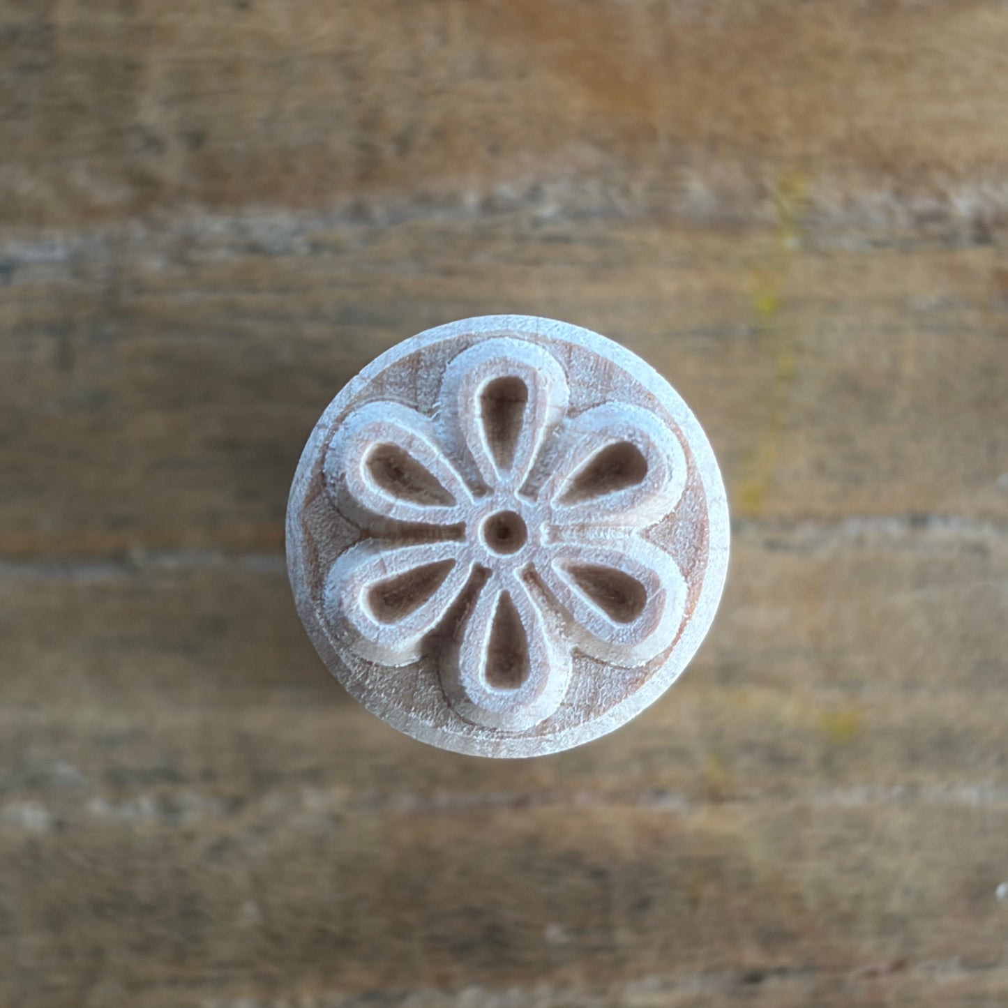 A round, light-colored wooden stamp or carving tool with a floral petal pattern, shown on a rustic wooden surface.