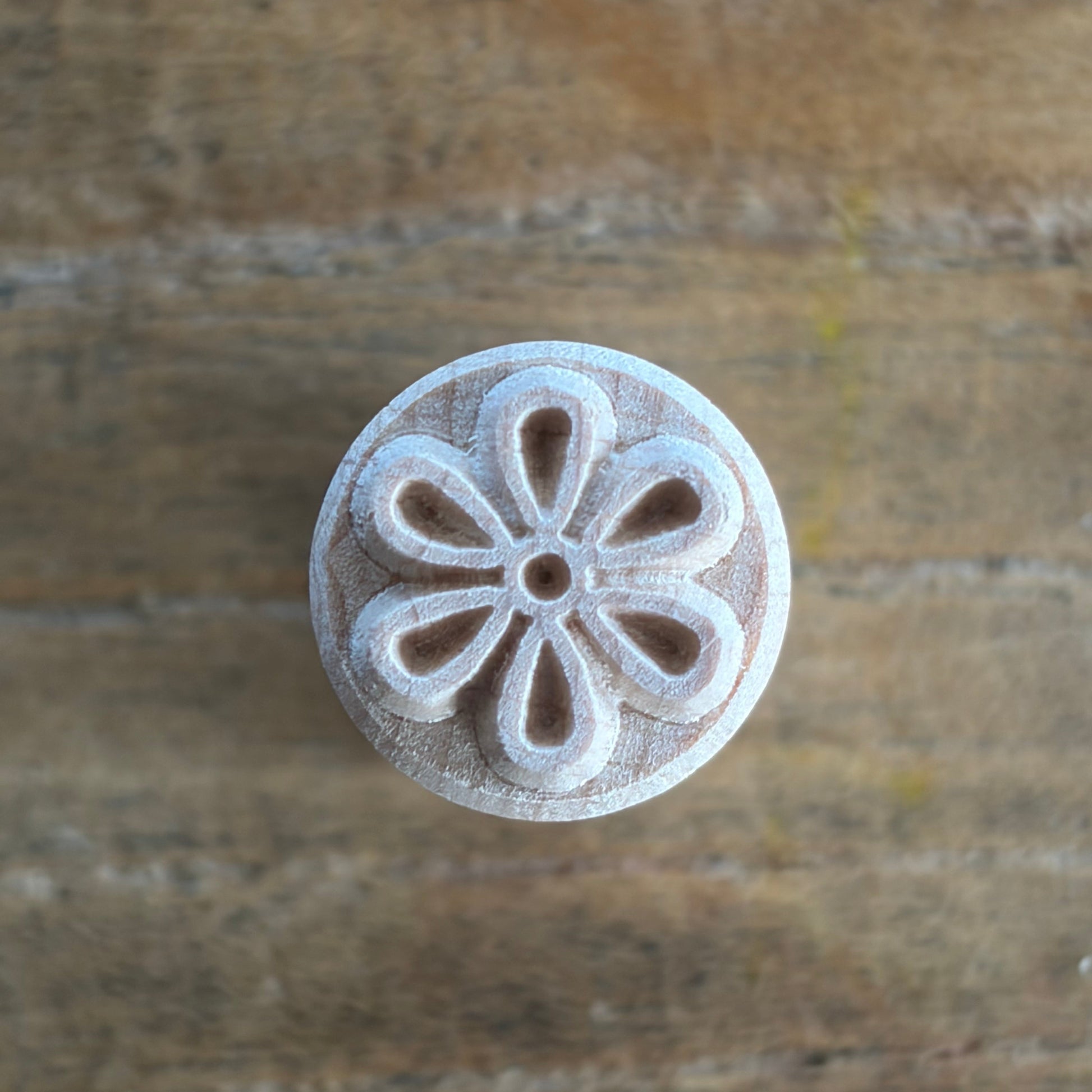A round, light-colored wooden stamp or carving tool with a floral petal pattern, shown on a rustic wooden surface.