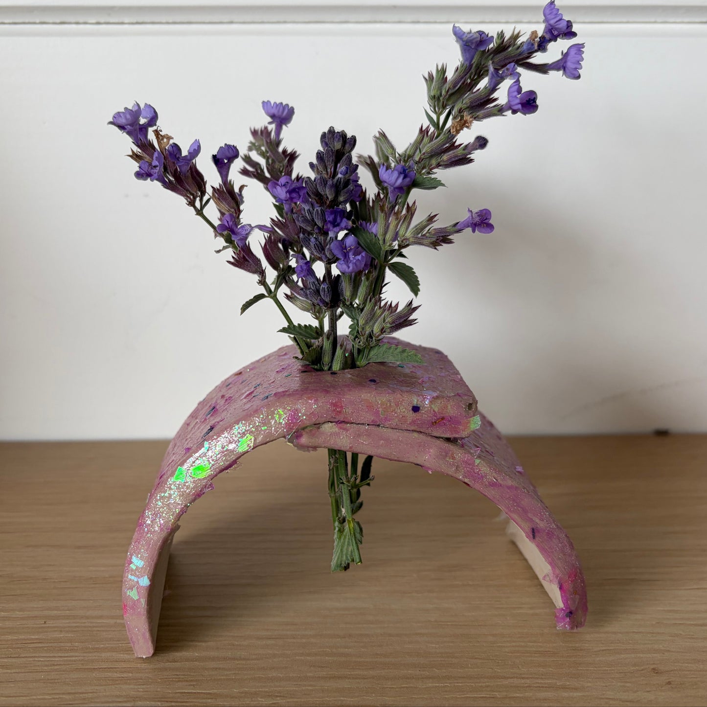 A small handmade pink arch-shaped planter made from painted air dry clay, holding a small bouquet of purple flowers, set on a wooden surface against a white wall.