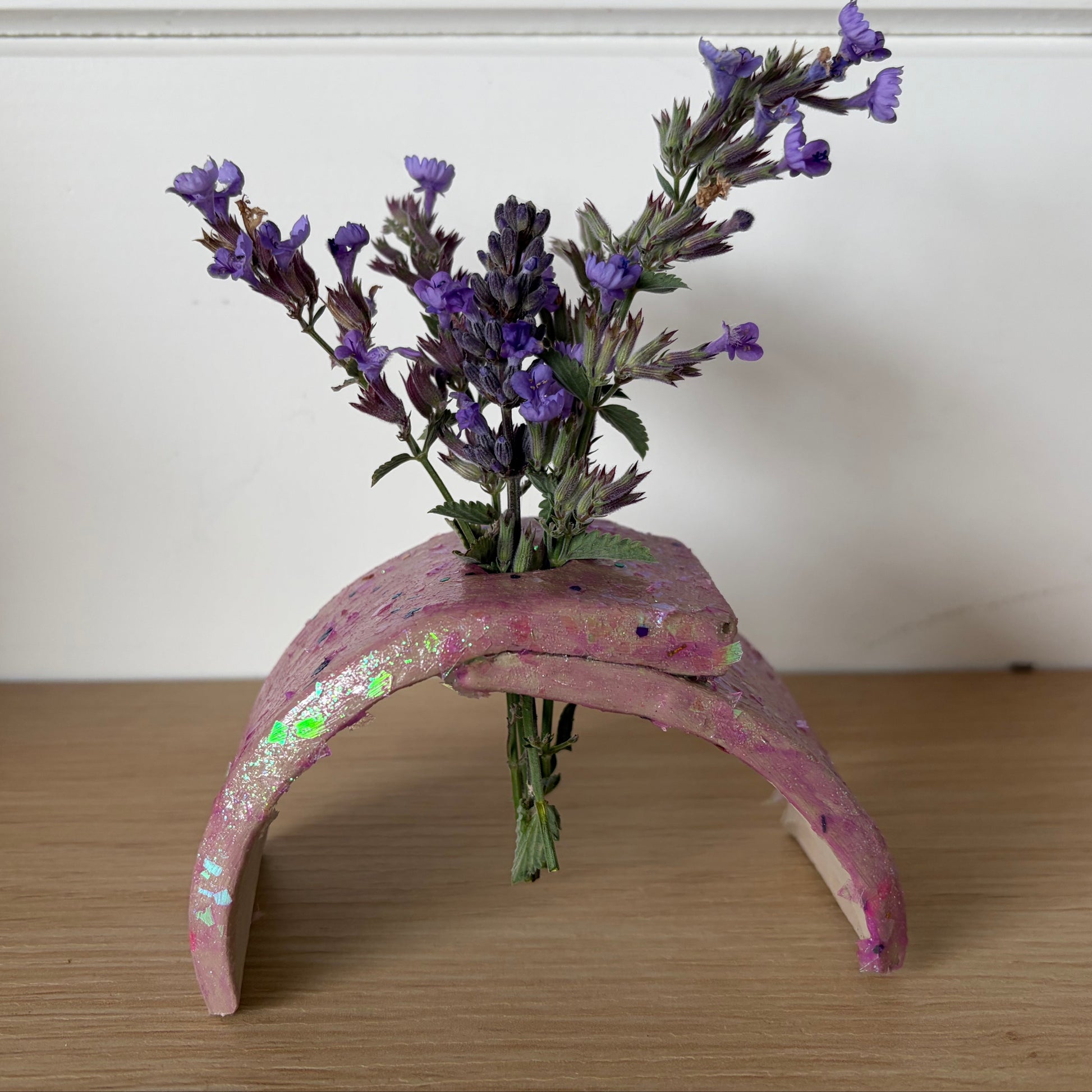 A small handmade pink arch-shaped planter made from painted air dry clay, holding a small bouquet of purple flowers, set on a wooden surface against a white wall.