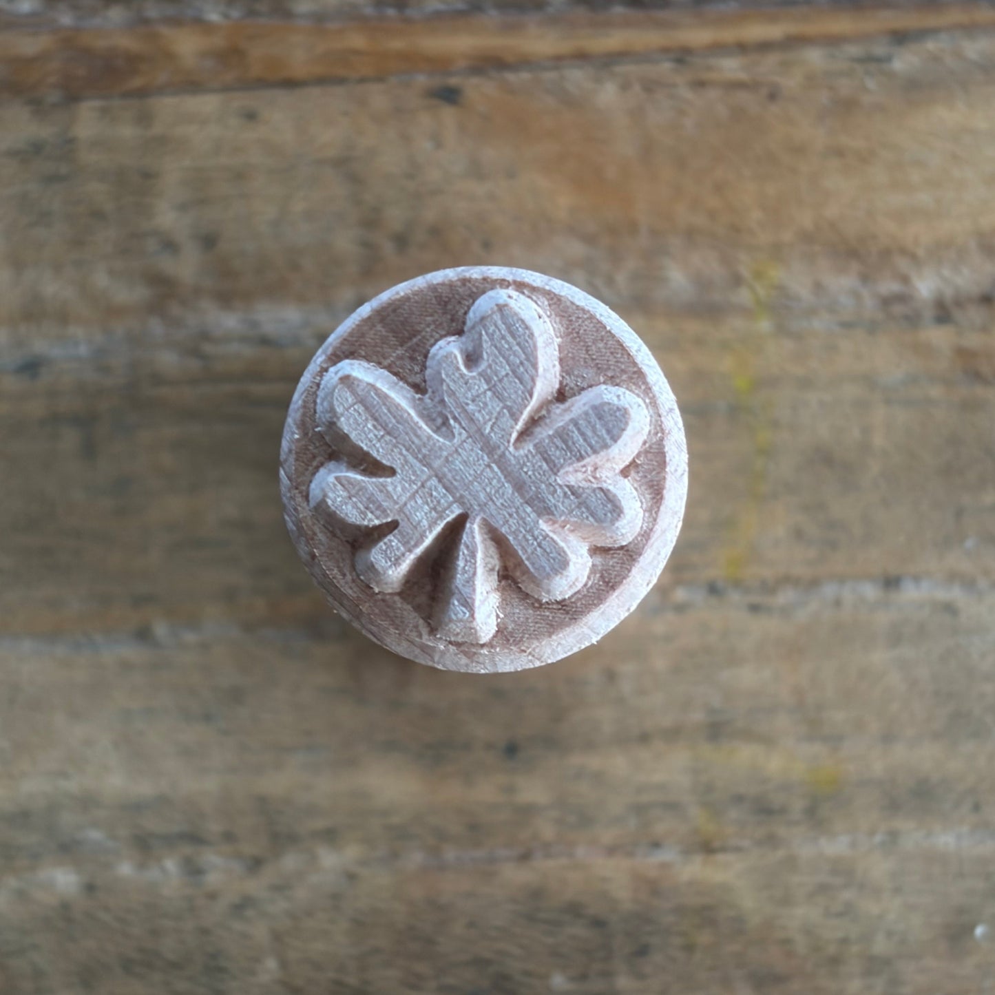 A round wooden stamp with a carved clover or leaf-like motif, shown from above on a rustic wooden surface.