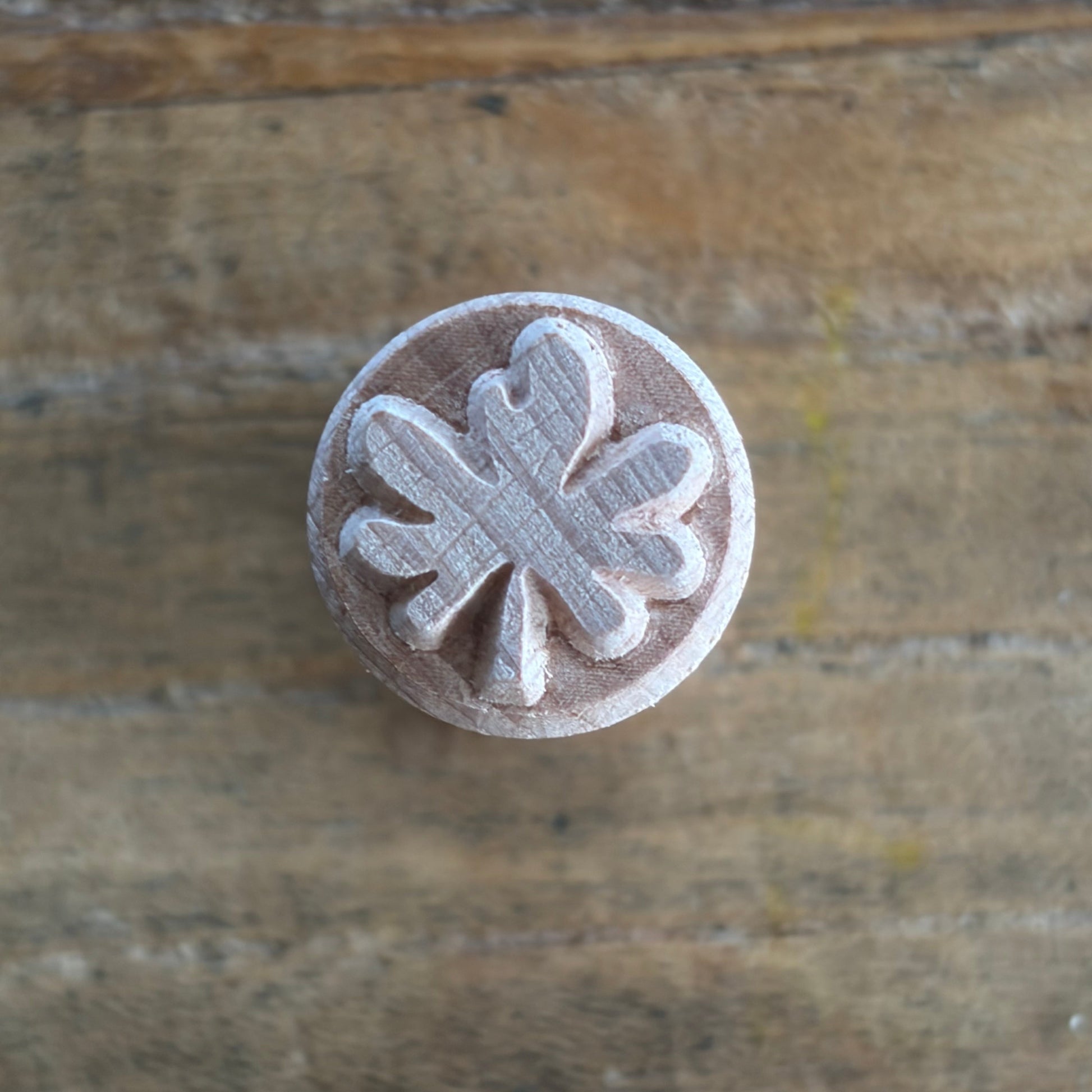 A round wooden stamp with a carved clover or leaf-like motif, shown from above on a rustic wooden surface.