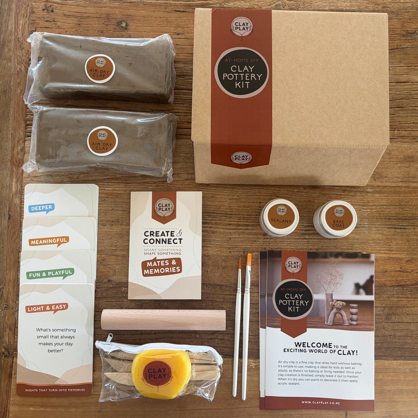 A flat-lay view of air dry pottery kit on a wooden table, containing clay, a pottery l, a clay roller, a yellow sponge, a paintbrush, tools and mates and memorie conversation cards