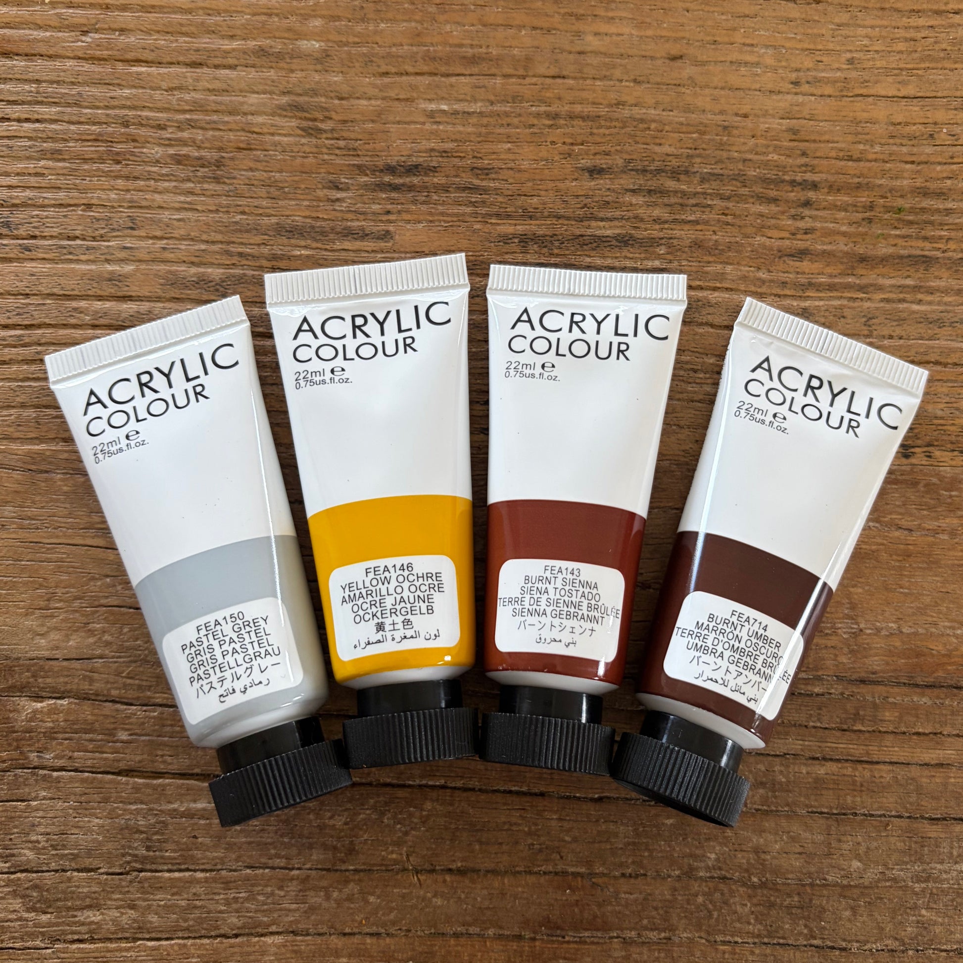 Four tubes of acrylic paint of natural colors are lined up on a wooden surface. The tubes are labeled 'ACRYLIC COLOUR' and show the colors: light gray, yellow, brown, and dark brown.