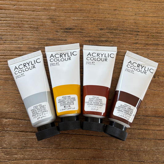 Four tubes of acrylic paint of natural colors are lined up on a wooden surface. The tubes are labeled 'ACRYLIC COLOUR' and show the colors: light gray, yellow, brown, and dark brown.