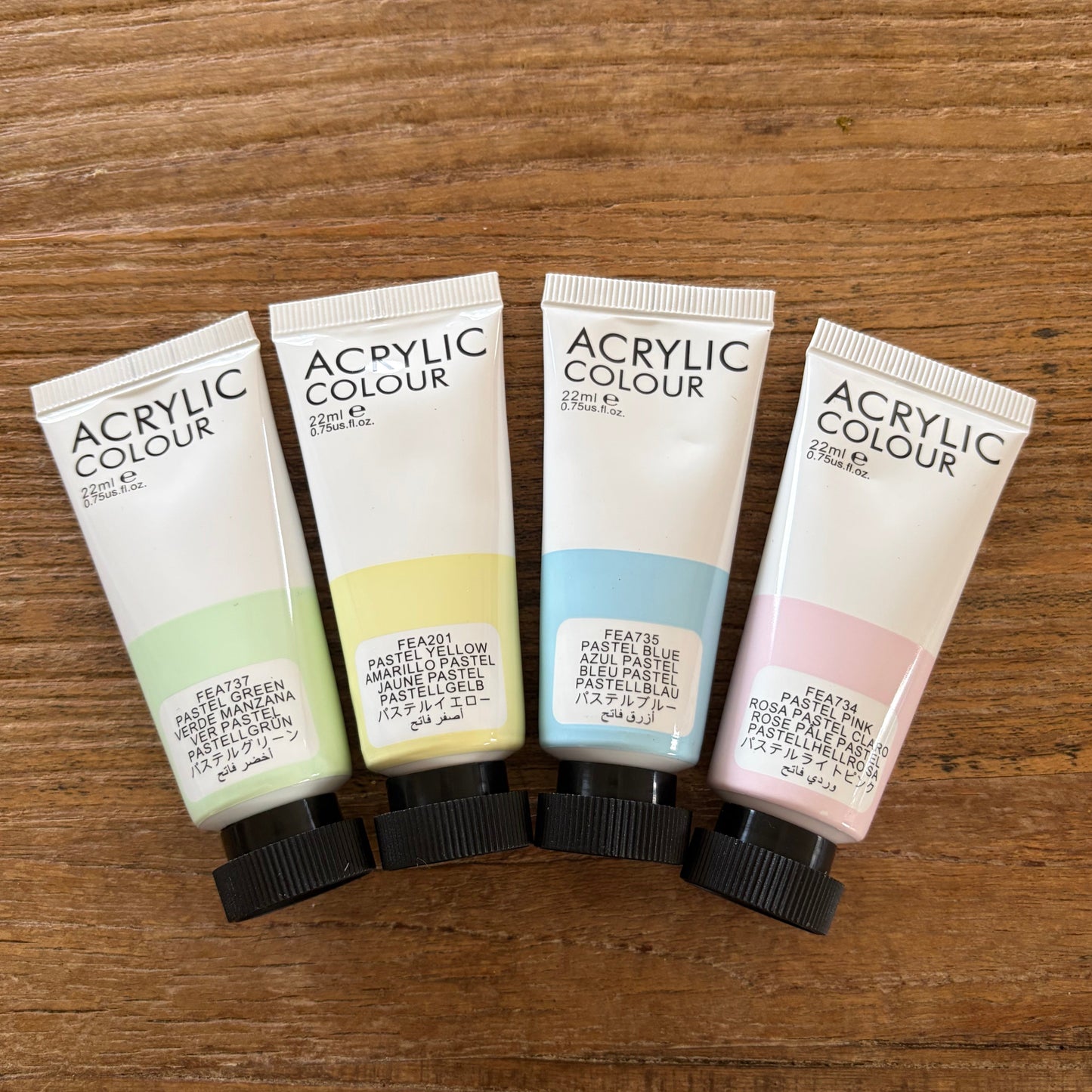 Four tubes of acrylic paint in pastel shades of green, yellow, light blue, and pink, arranged diagonally on a brown wooden surface.