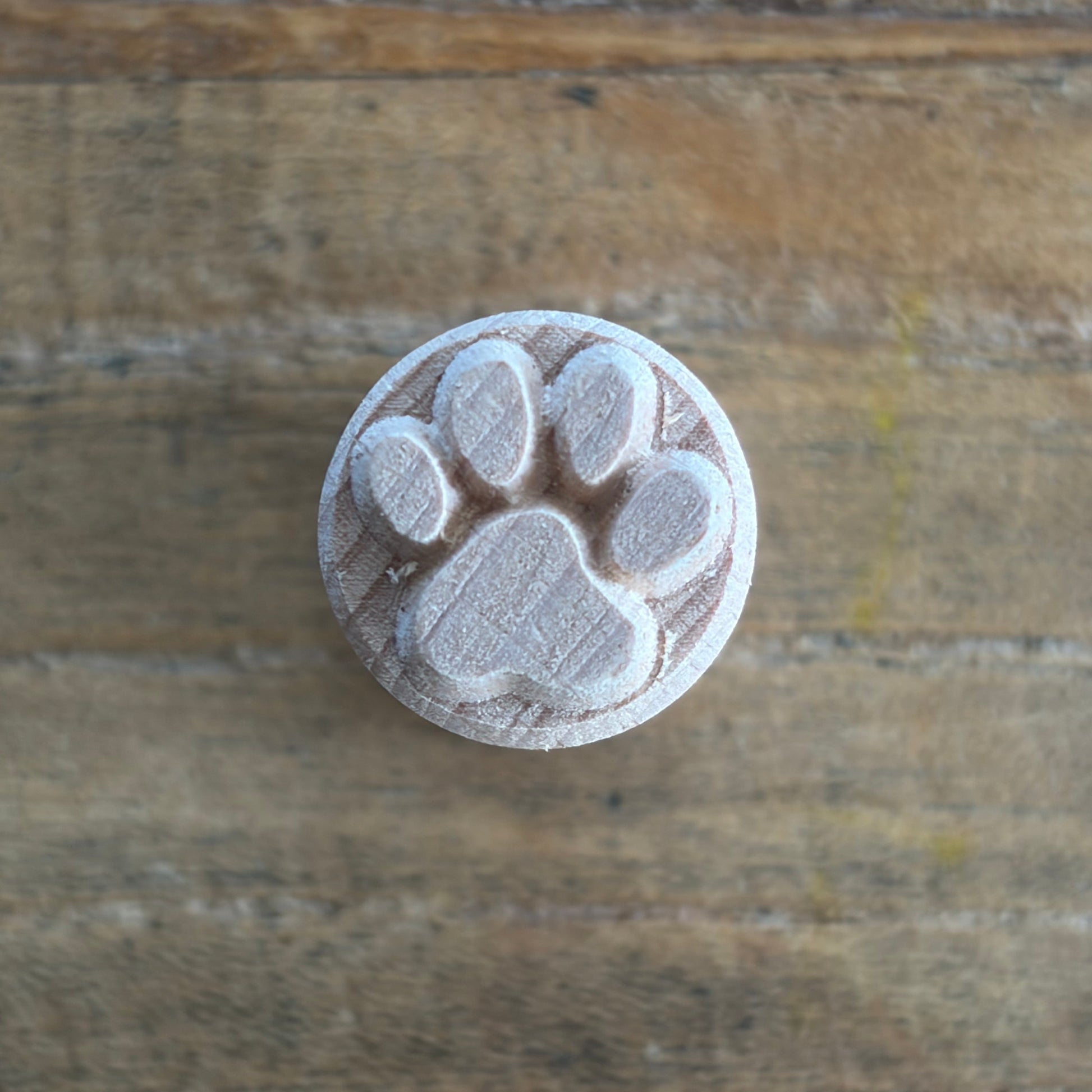 A round wooden stamp featuring an embossed paw print design, shown on a rustic wooden surface.