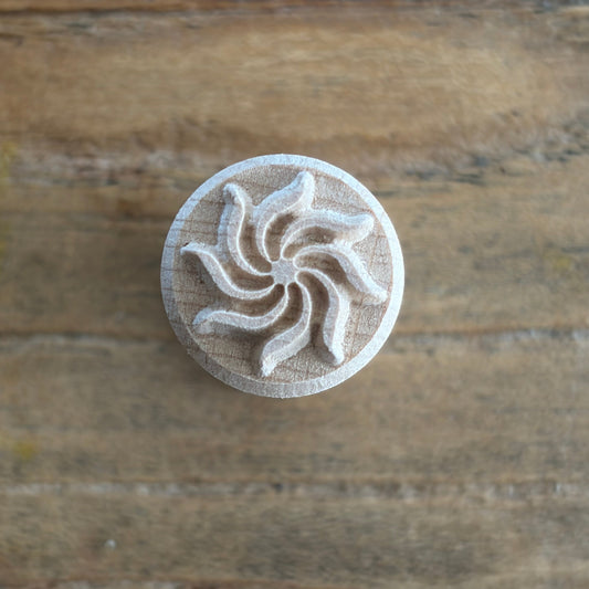 A round wooden stamp with a carved spiral or windmill-like design, photographed from above on a rustic wooden surface.